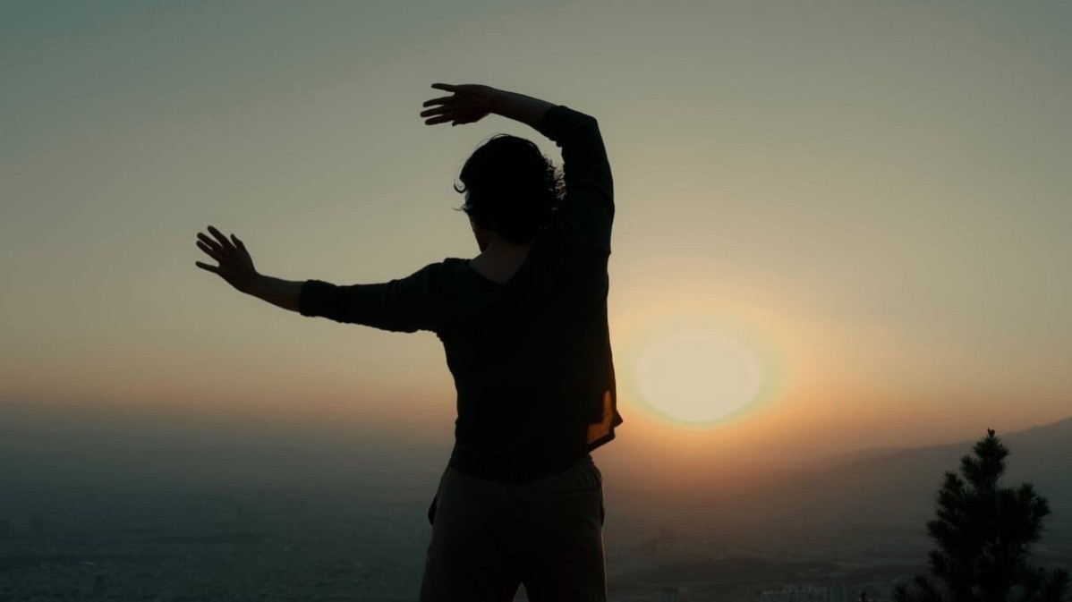 Silhouette einer Person mit ausgestreckten Armen vor Sonnenuntergang über einer Landschaft