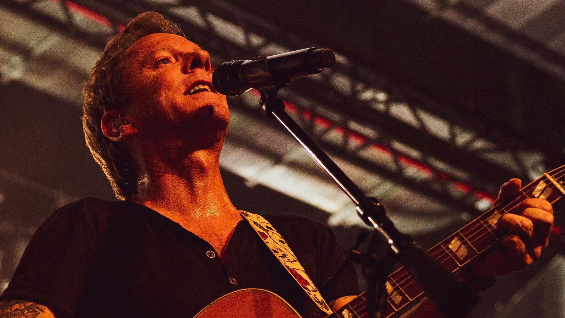 Kiefer Sutherland singt live auf der Bühne, hält eine Gitarre und steht vor einem Mikrofon, warmes Bühnenlicht und Konzertatmosphäre.