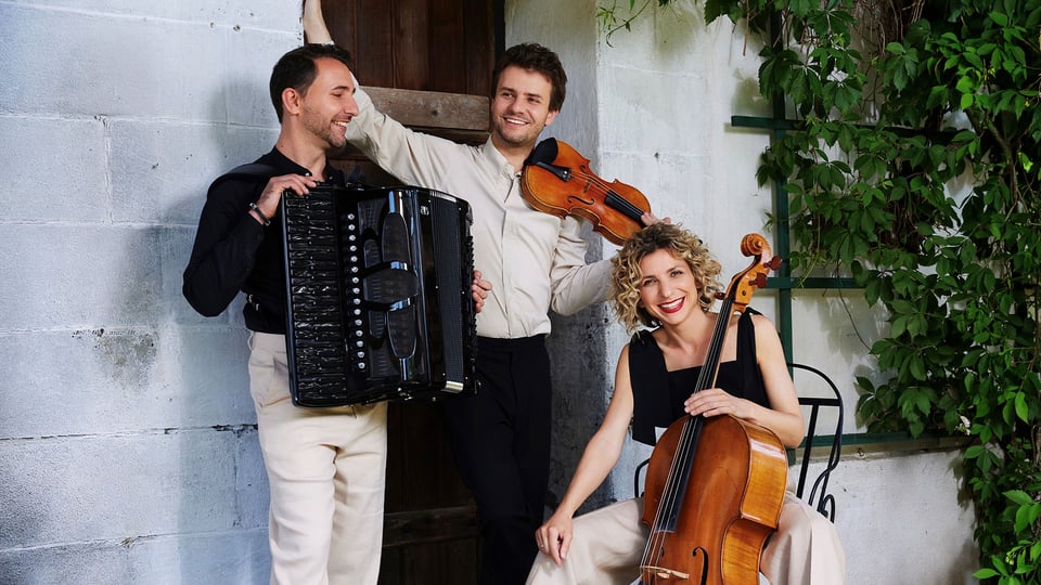 Bandfoto Trio Wellcaru mit drei Personen vor einer weßen Ziegelwand mit Holztür, eine Person mit Akkordeon eine mit Geige und eine mit Cello daneben rankende Pflanzen