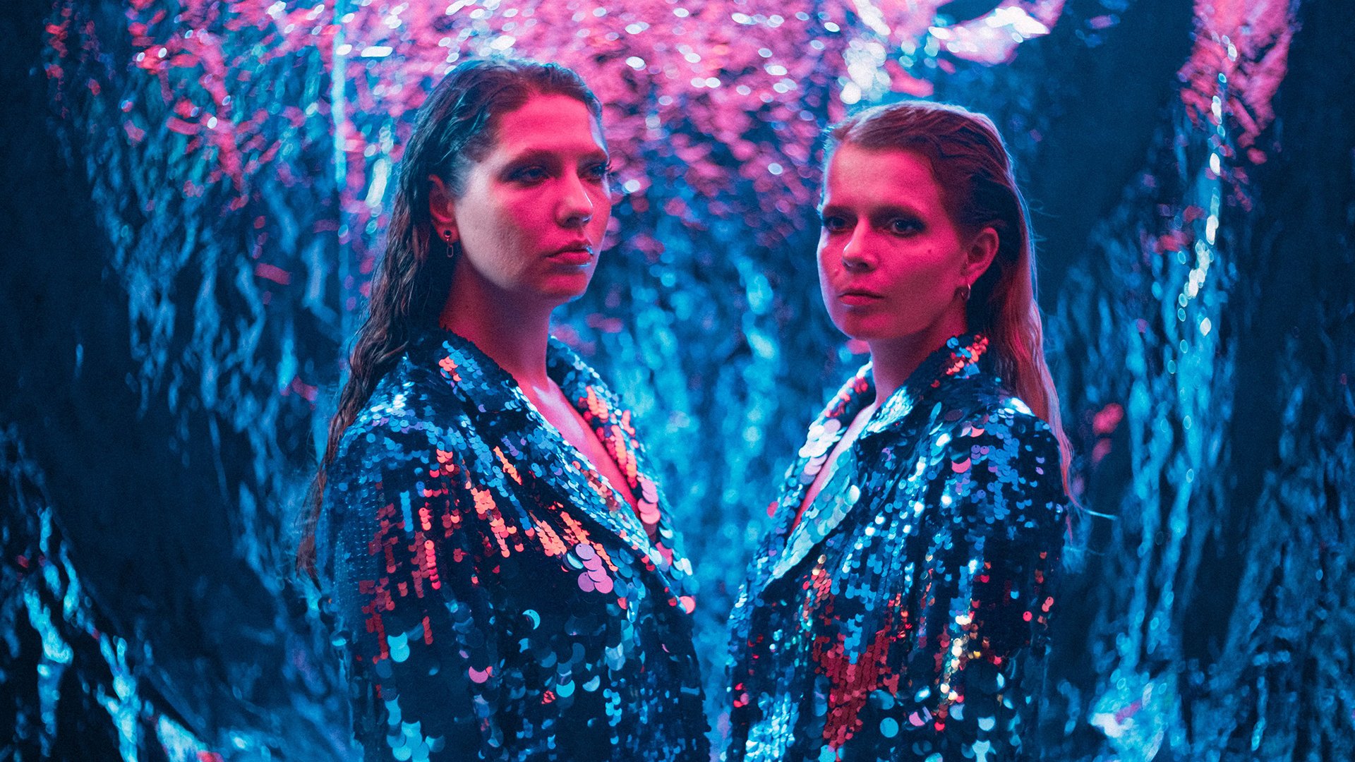 Zwei Frauen stehen sich gegenüber und stehen gekleidet in Pailletten vor einem reflektierenden Hintergrund