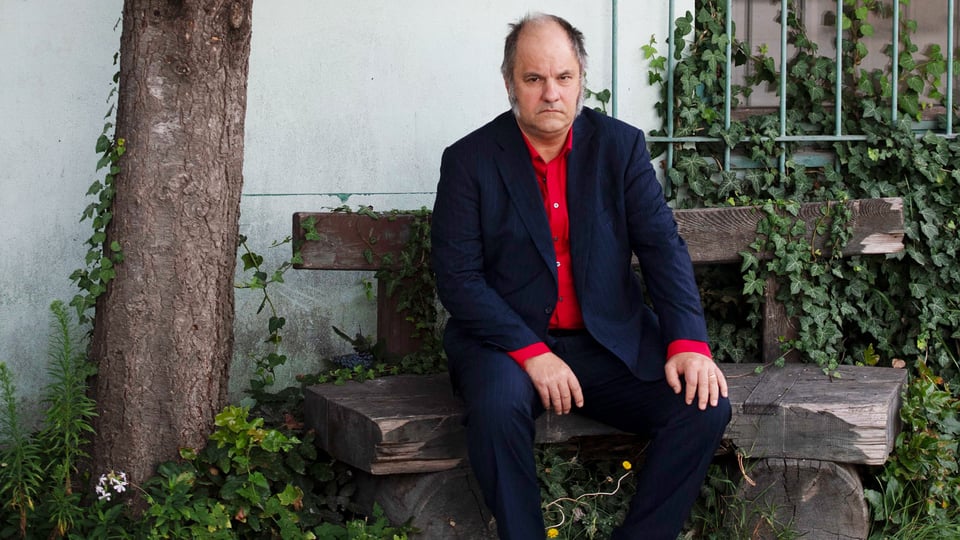 Foto für das Event Mathias Egersdörfer – langsam mit dem Künstler, der auf einer Holzbank sitzt und ein rotes Hemd trägt