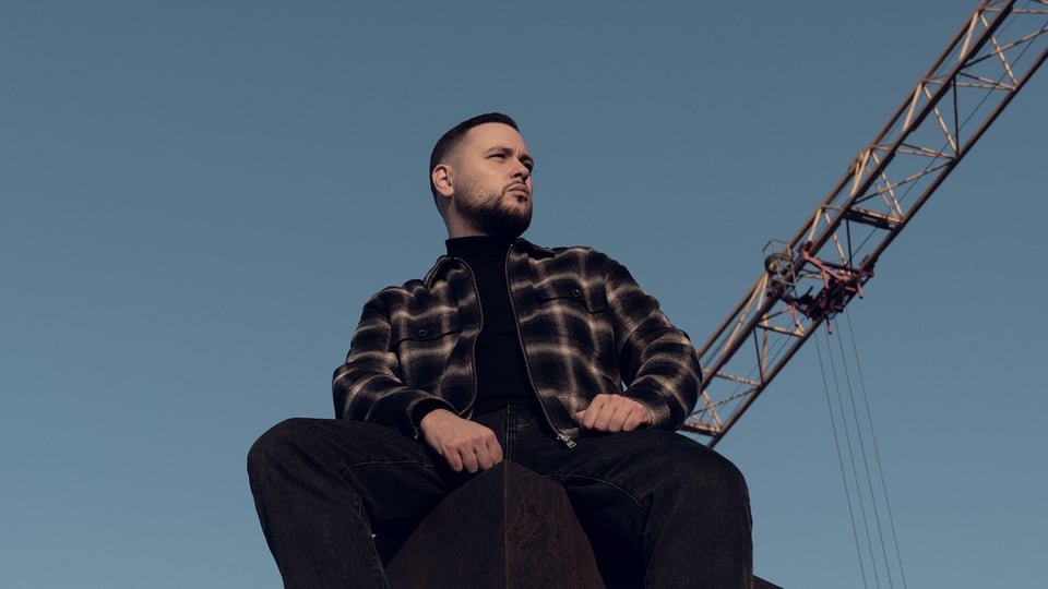 Der Künstler PTK sitzt erhöht vor blauem Himmel, trägt dunkle Kleidung und blickt seitlich in die Ferne, im Hintergrund ragt ein Baukran ins Bild.