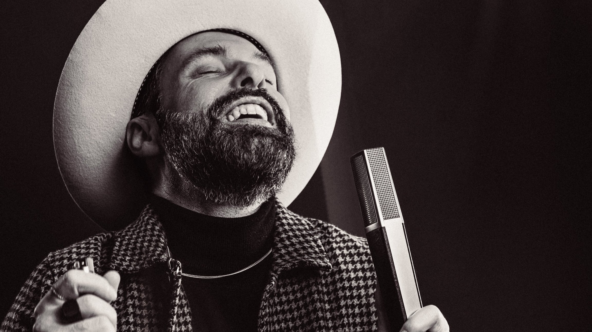 Schwarz‑weißes Porträt von San2 mit Bart und hellem Hut, hält ein Mikrofon in der Hand; Mund geöffnet, kariertes Jackett, dunkler Hintergrund mit Bühnenlicht.