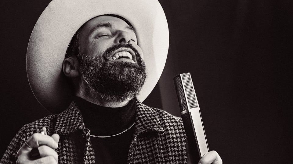 Schwarz‑weißes Porträt von San2 mit Bart und hellem Hut, hält ein Mikrofon in der Hand; Mund geöffnet, kariertes Jackett, dunkler Hintergrund mit Bühnenlicht.