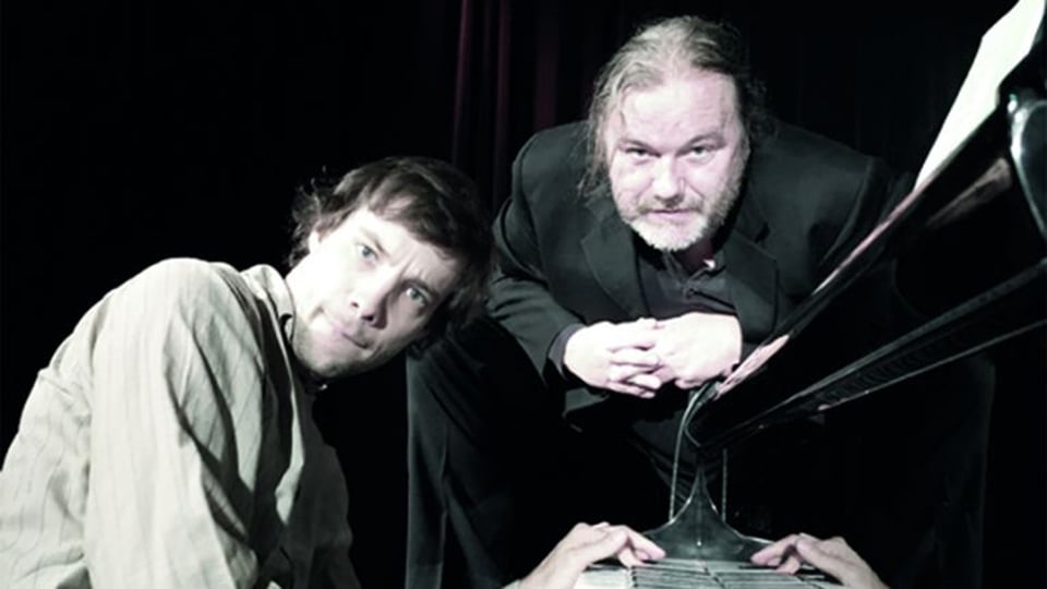 Foto für die Veranstaltung ‚Im Auftrag des Herrn‘ mit den beiden Künstlern, wobei einer von ihnen am Klavier sitzt.