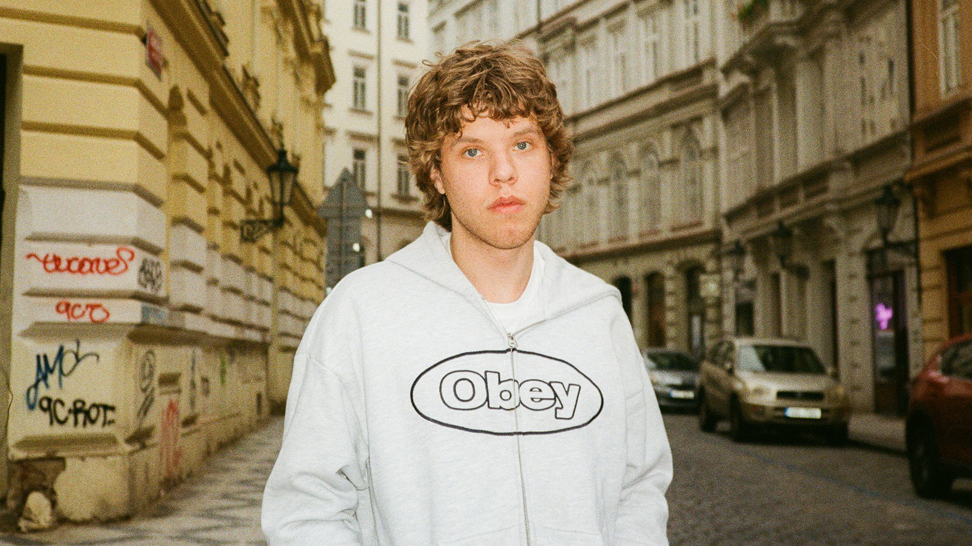 Der Künstler Zartmann mit ernstem Blick in weißem Sweatshirt mit der Aufschrift Obey steht vor einer Häuserzeile aus altertümlichen Gebäuden und Graffitian einer Wand