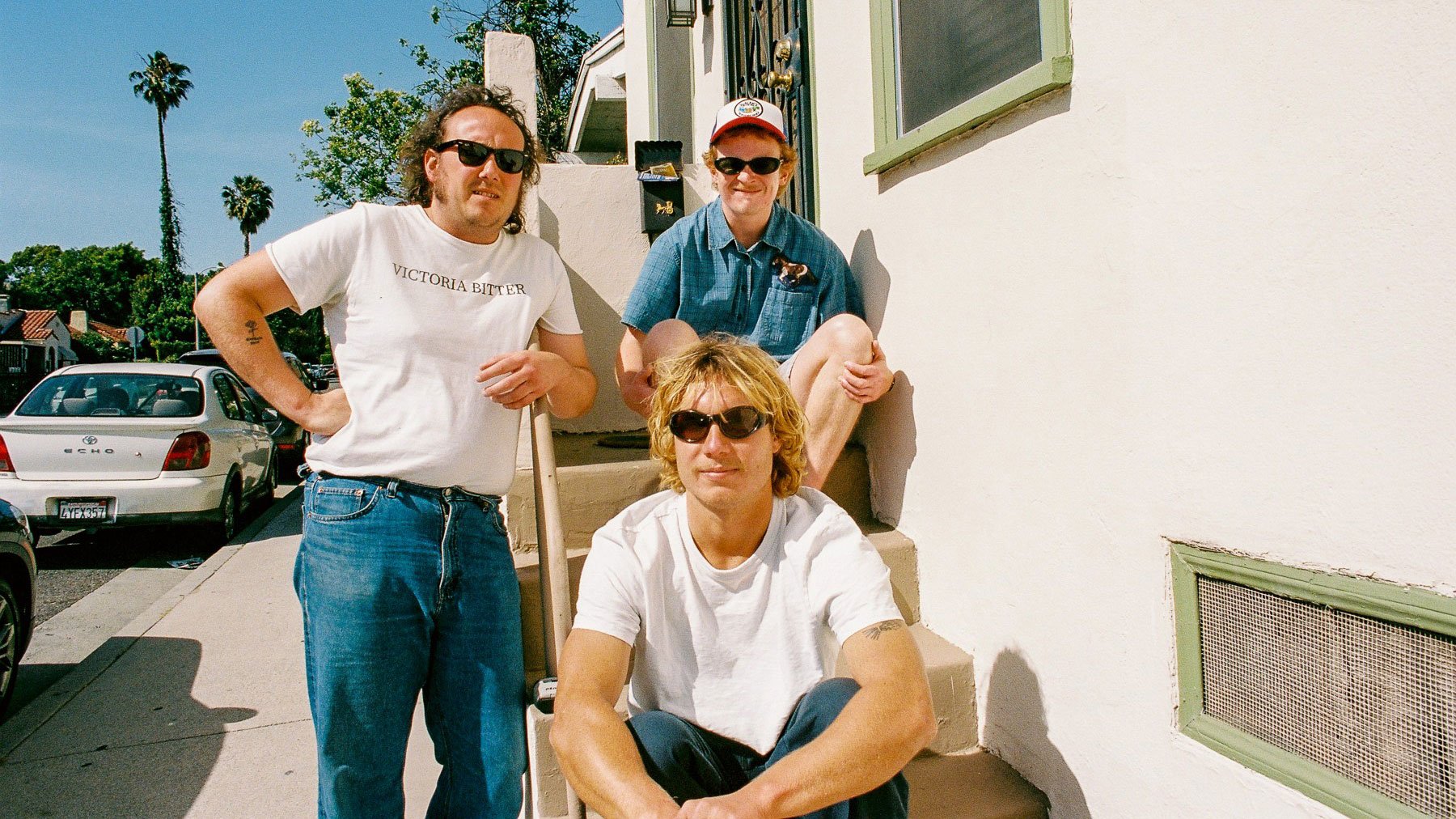 Drei Mitglieder der Band The Chats sitzen und stehen auf der Treppe eines Wohnhauses in einer sonnigen Wohnstraße. Sonnenbrillen, helle T Shirts, Jeans und eine Baseballkappe prägen die Aufnahme. Palmen, geparkte Autos und Hausfassaden bilden den Hintergrund.