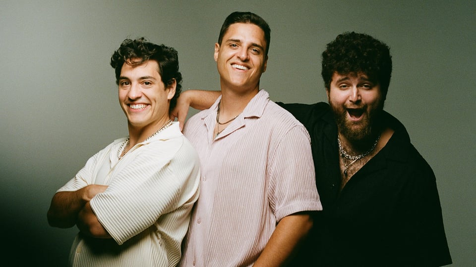 Gruppenfoto von OCT mit drei Personen vor grauem Hintergrund, eng nebeneinander stehend und mit hellen und dunklen Hemden.