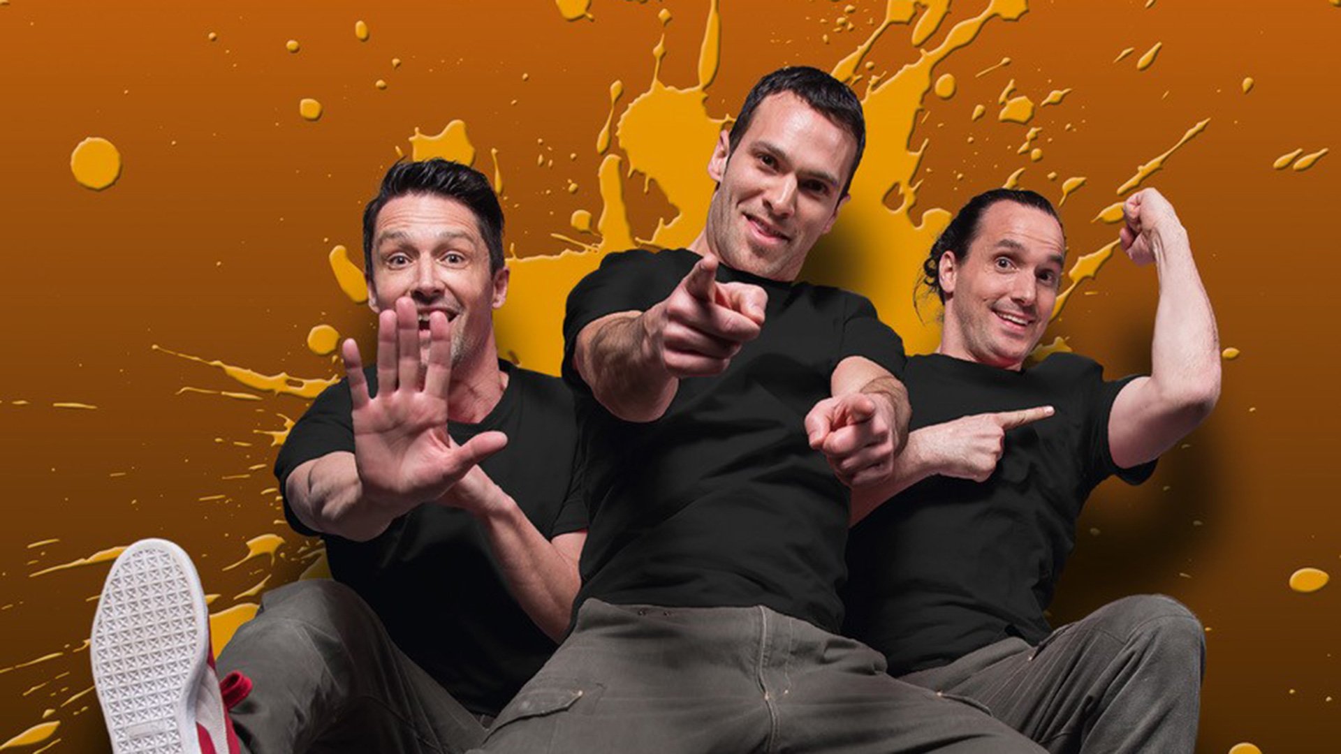 Foto zu „Starbugs Comedy – JUMP! Reloaded“ mit drei Männern in schwarzen T-Shirts vor orangefarbenem Hintergrund, in dynamischen Posen.