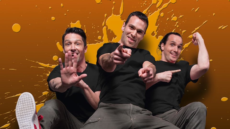 Foto zu „Starbugs Comedy – JUMP! Reloaded“ mit drei Männern in schwarzen T-Shirts vor orangefarbenem Hintergrund, in dynamischen Posen.