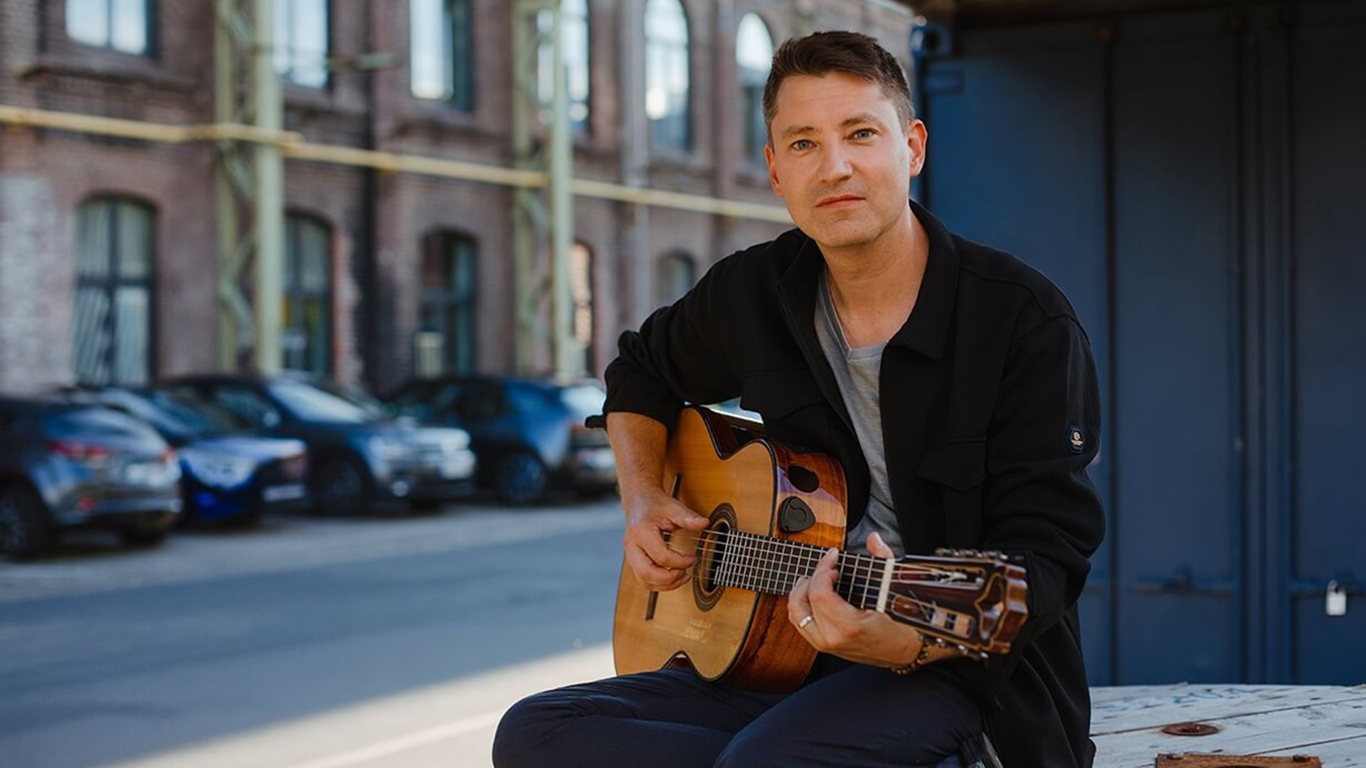 Daniel Stelter sitzend mit Gitarre in schwarzem Hemd, im Hintergrund eine Straßenszenen mit Hausfassade und parkenden Autos