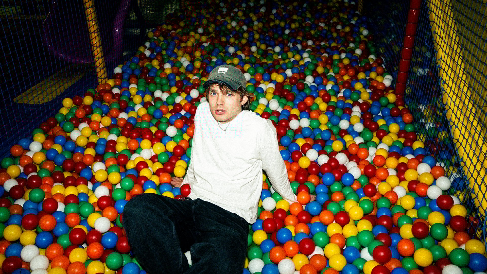 Jaschu sitzt in einem bunten Bällebad aus Plastikkugeln und blickt nach oben zur Kamera mit Netzen im Hintergrund.