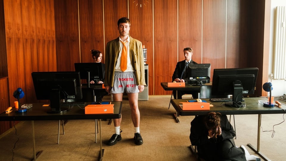 Büroszene mit mehreren Schreibtischen und Computern; eine Person steht zentral im Sakko, Krawatte und Shorts, andere sitzen oder hocken an Arbeitsplätzen vor holzvertäfelter Wand