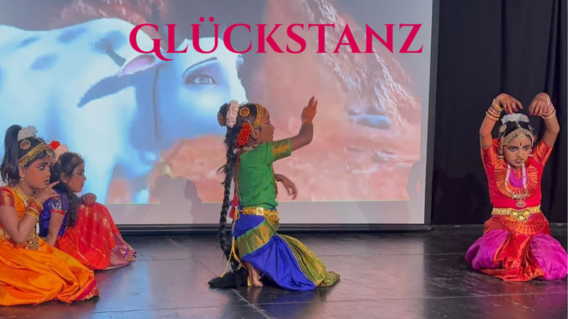 Bühnenfoto Glückstanz mit Kindern in farbenfrohen Tanzkostümen bei einer klassischen indischen Tanzaufführung vor projiziertem Hintergrund mit dem Schriftzug GLÜCKSTANZ