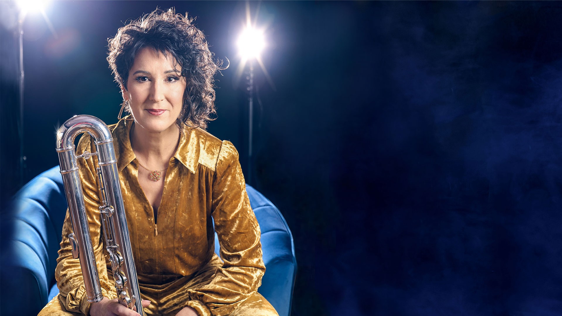 Isabelle Bodenseh auf einem blauen Sessel. Sie trägt goldfarbene Kleidung und hält eine silberne Bassklarinette. Dunkelblauer Hintergrund mit zwei hellen Scheinwerfern.