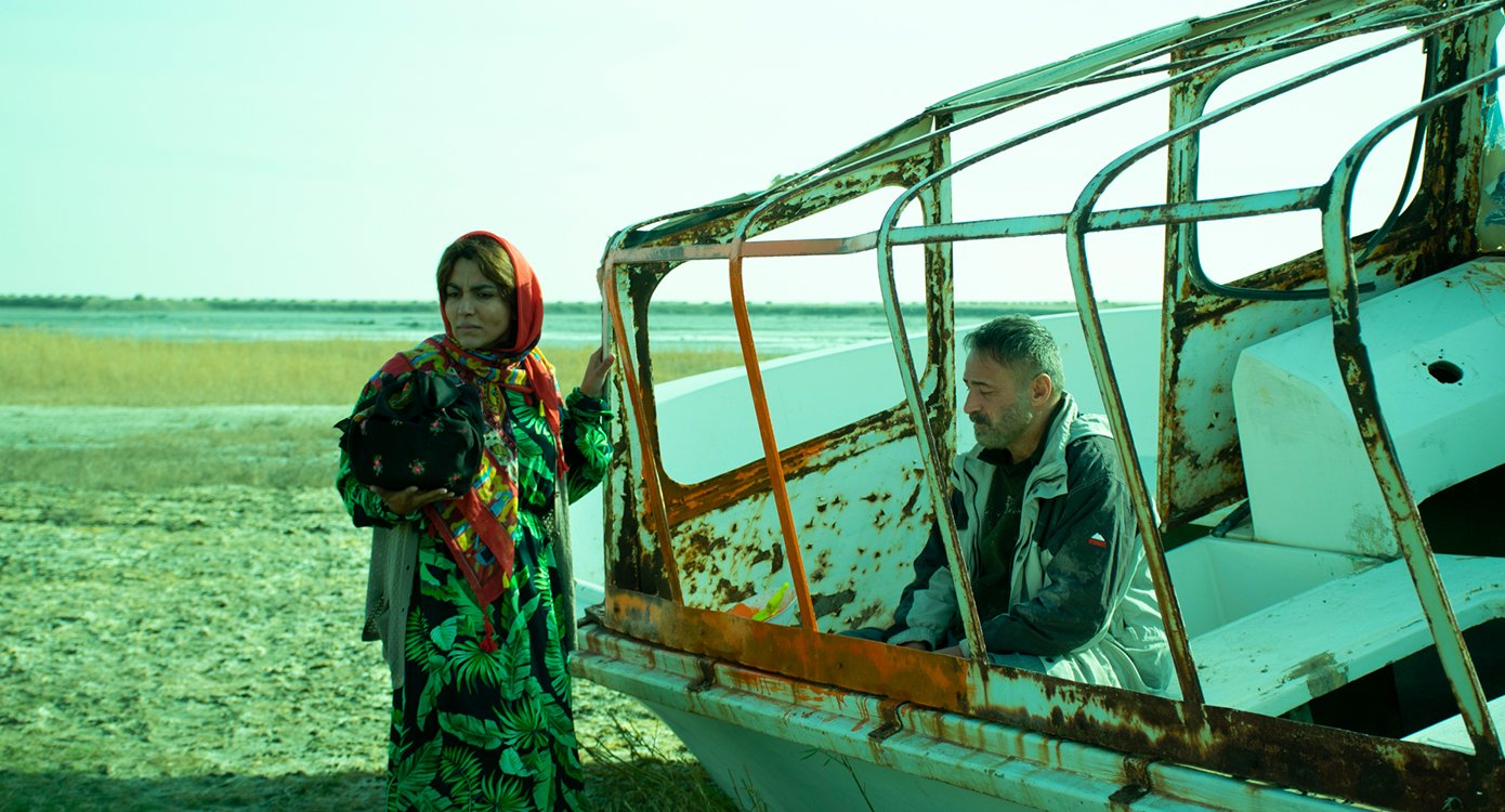 eine Dame mit Kopftuch und ein Mann, der auf einem verfallenen Boot am Strand sitzt