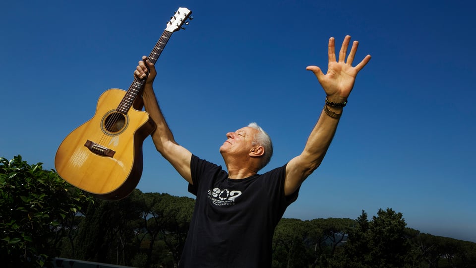 Eventmotiv von Tommy Emmanuel: stehende Person im Freien vor blauem Himmel, hält eine akustische Gitarre erhoben in einer Hand, die andere Hand ist nach oben ausgestreckt. Dunkles T‑Shirt, Tageslicht; Motiv zur Live‑Konzertankündigung des Gitarristen Tommy Emmanuel.