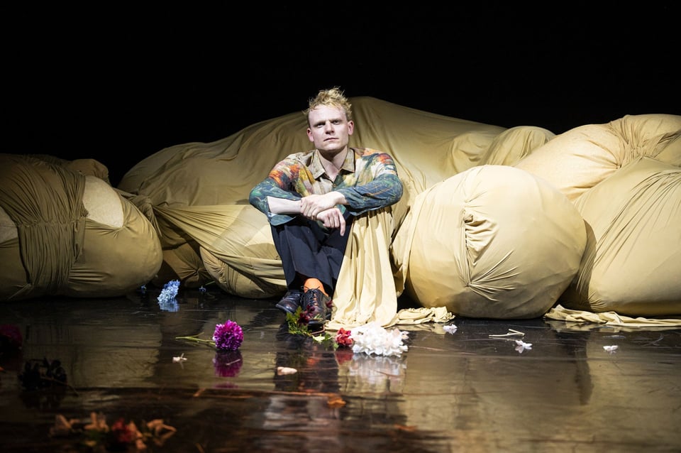 Sitzende Person auf einer Bühne zwischen großen, beigefarbenen Stoffformen, umgeben von verstreuten Blumen, vor dunklem Hintergrund.