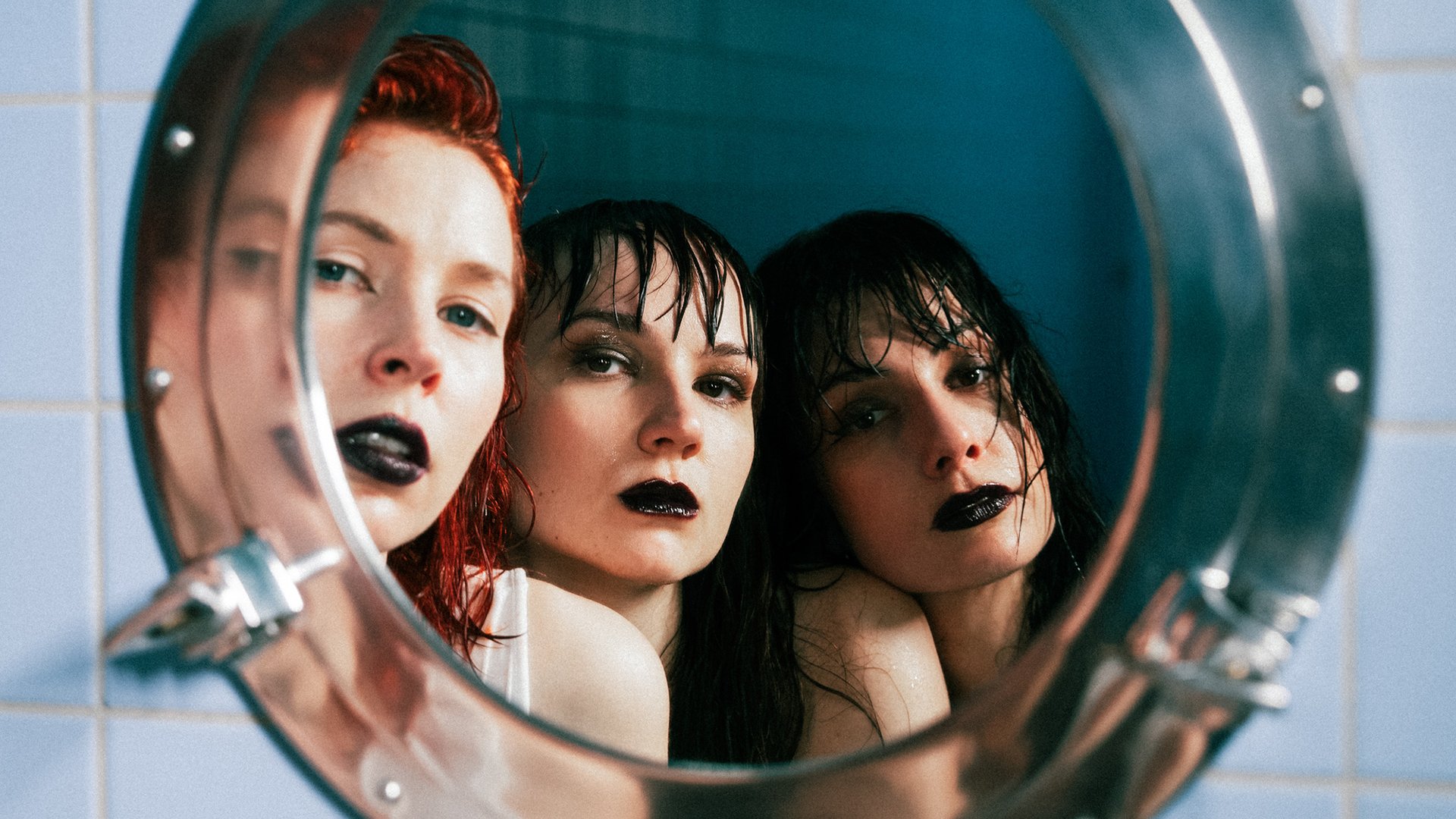 My Ugly Clementine Bandfoto: Drei Frauen mit dunklem Make-up blicken in einen runden Spiegel, nasse Haare, Badezimmer-Setting mit kühlen Farbtönen, direkter Blick in die Kamera.