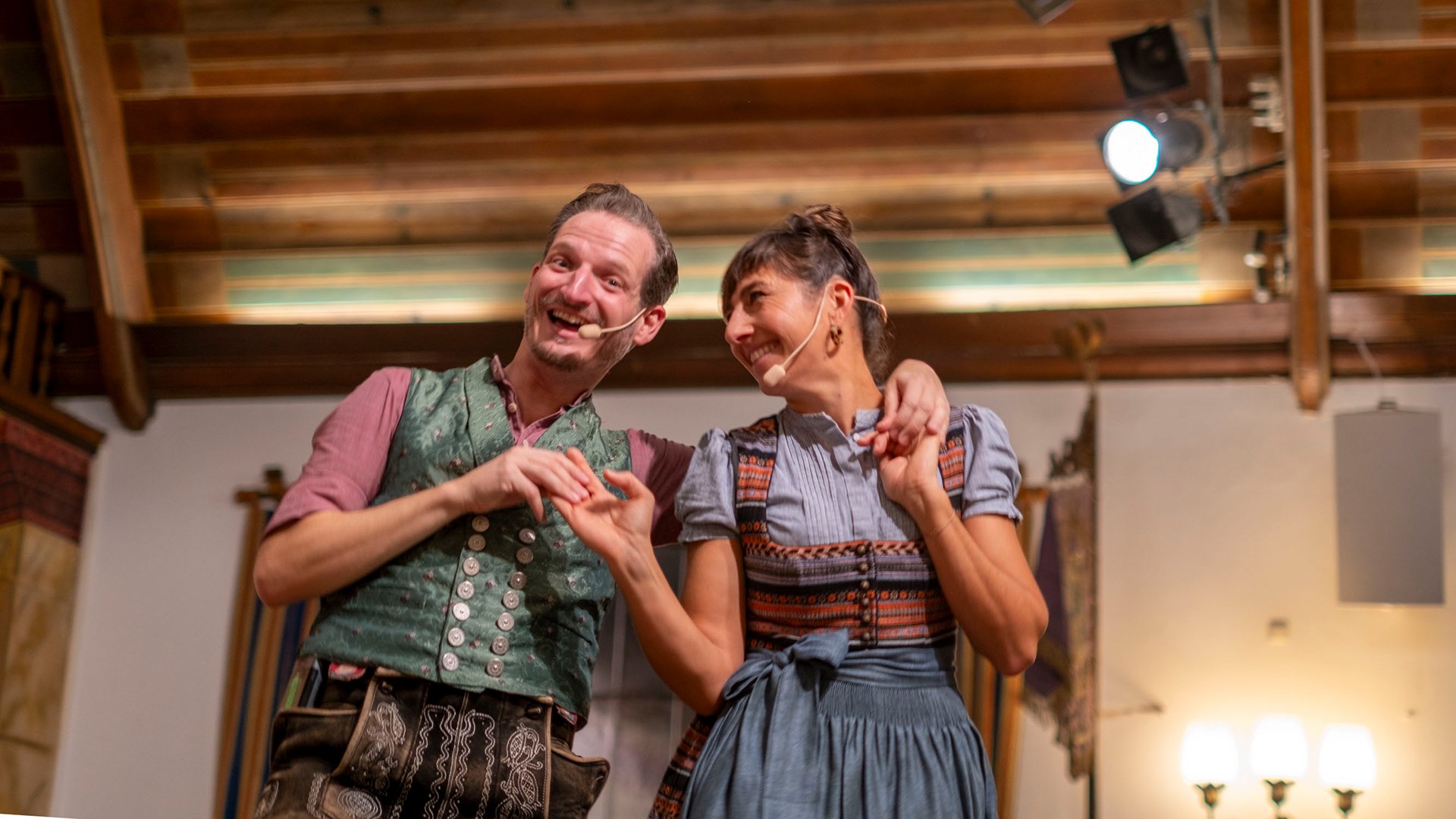 Bühnenfoto vom Kathreintanz mit zwei Personen in bayerischer Tracht, eine in grün gemusterter Weste und Lederhose, die andere im blauen Dirndl, auf einer Bühne mit Headsetmikrofonen und warm beleuchtetem Saal im Hintergrund