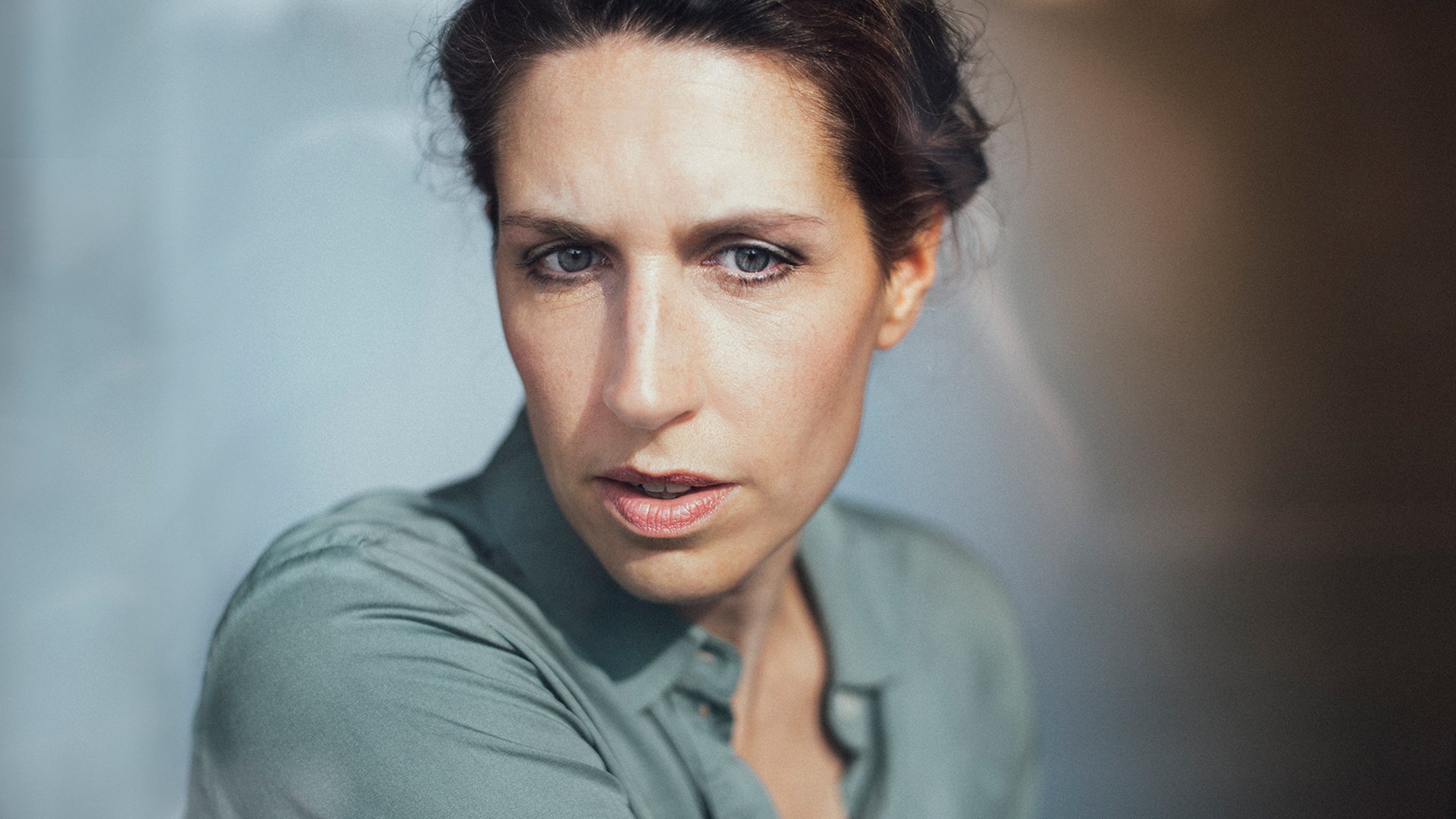 Portrait von Christine Prayon in türkiser Bluse vor hellblau-hellbraunem Hintergrund mit Farbverlauf