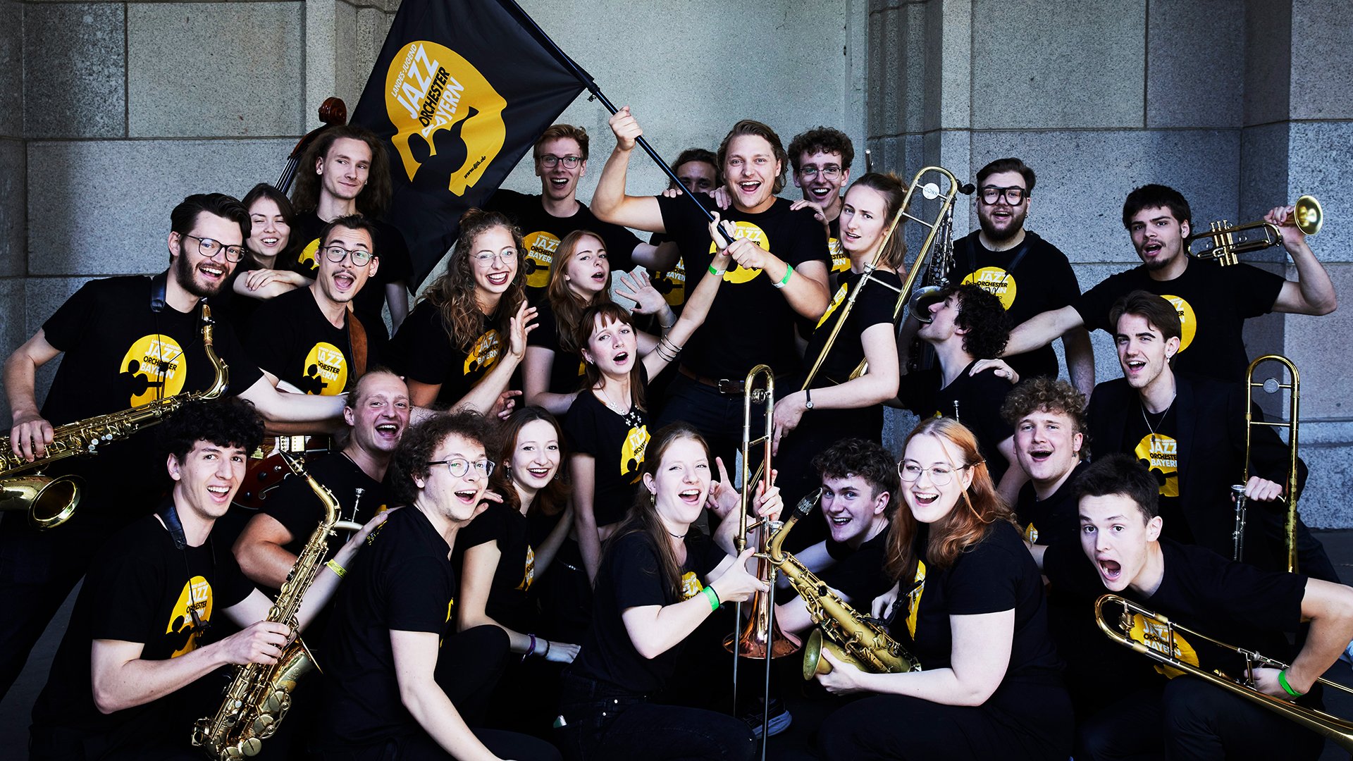 Eine große Gruppe junger Musikerinnen und Musiker posiert mit Saxofonen, Trompeten und Posaunen. In der Mitte wird eine Fahne des Landesjugendjazzorchesters Bayern hochgehalten. Die Stimmung ist fröhlich und ausgelassen.
