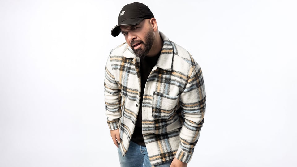 Pressefoto des Comedians Abdel Boudii vor hellem Hintergrund, mit Kappe und kariertem Hemd.