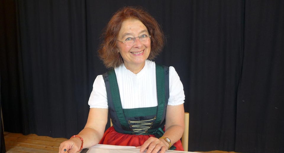 Foto einer Frau, die auf einem Stuhl sitzt und sich auf einen Tisch lehnt, in traditioneller bayerischer Tracht vor schwarzem Hintergrund.