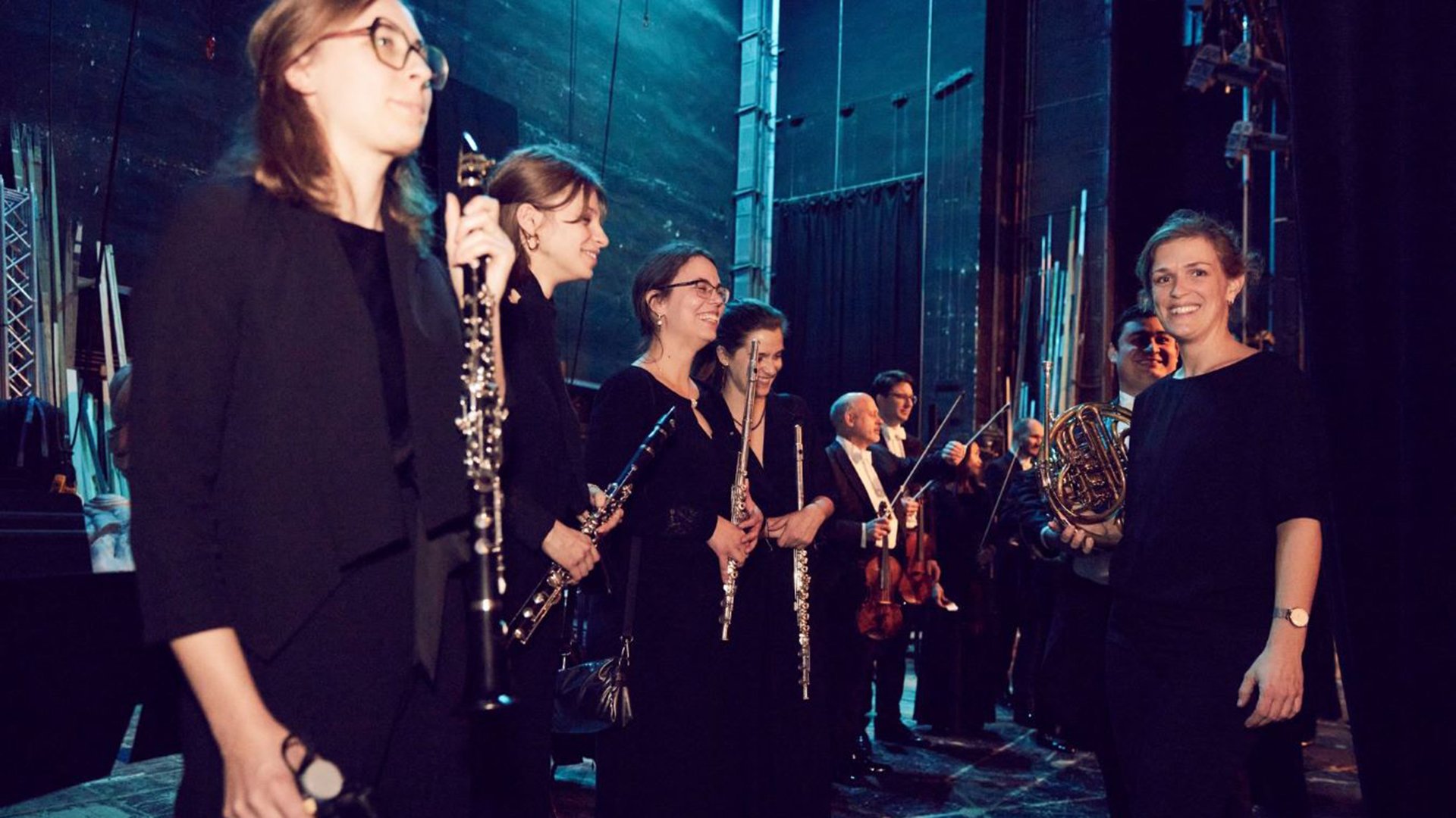 Mitglieder eines Orchesters stehen auf der Bühne und halten ihre Instrumente, darunter Holzbläser, Horn und Streicher; sie blicken seitlich zur Kamera.