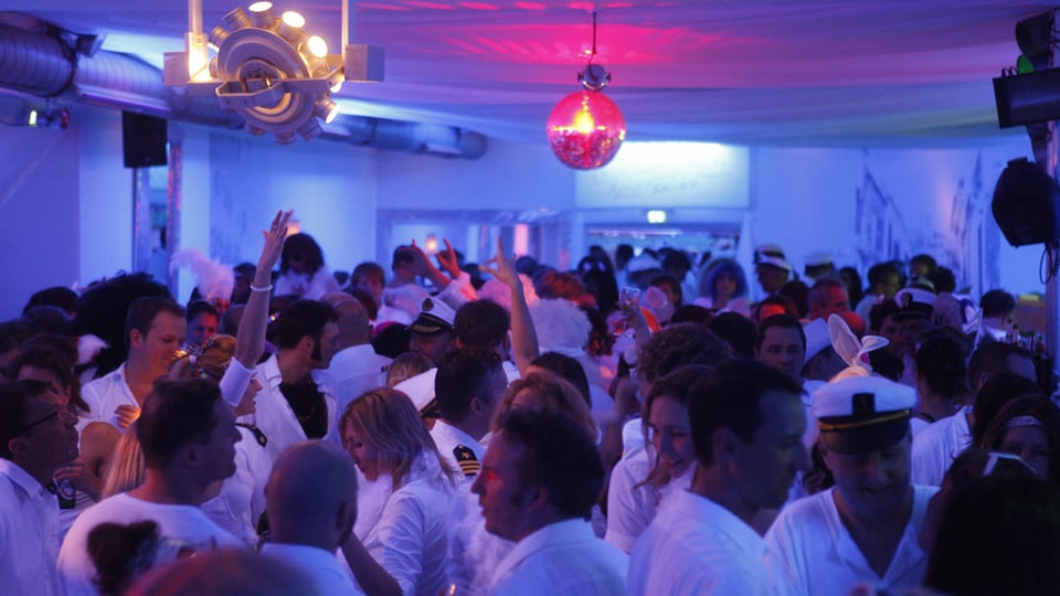 Weiß gekleidete Personen auf einer Tanzfläche in Schwarzlicht, an der Decke eine Diskokugel