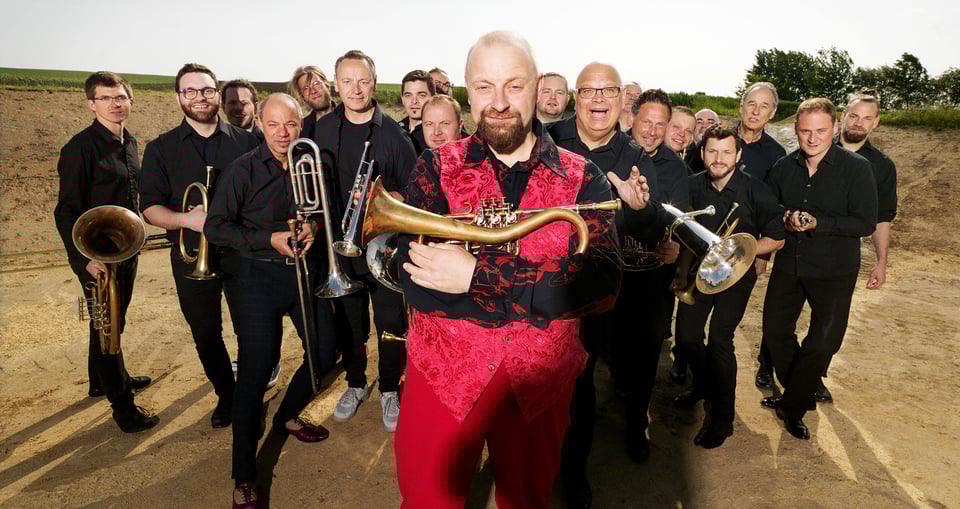 Foto der Gruppe mit allen schwarz gekleideten Musikern, außer einem rot gekleideten, mit Instrumenten