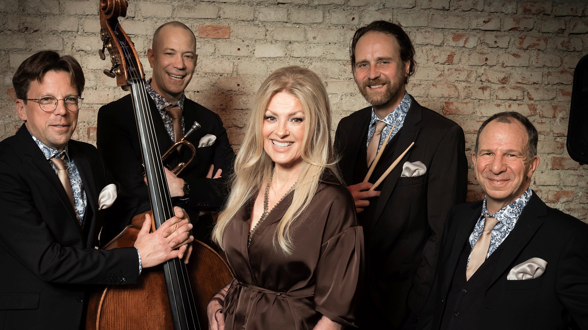 Gruppenfoto von Jasmin Bayer und Band vor einer Backsteinwand. Die Gruppe trägt dunkle Anzüge mit gemusterten Hemden, während ein Kontrabass, eine Trompete und Drumsticks als Instrumentelemente im Bild zu sehen sind.