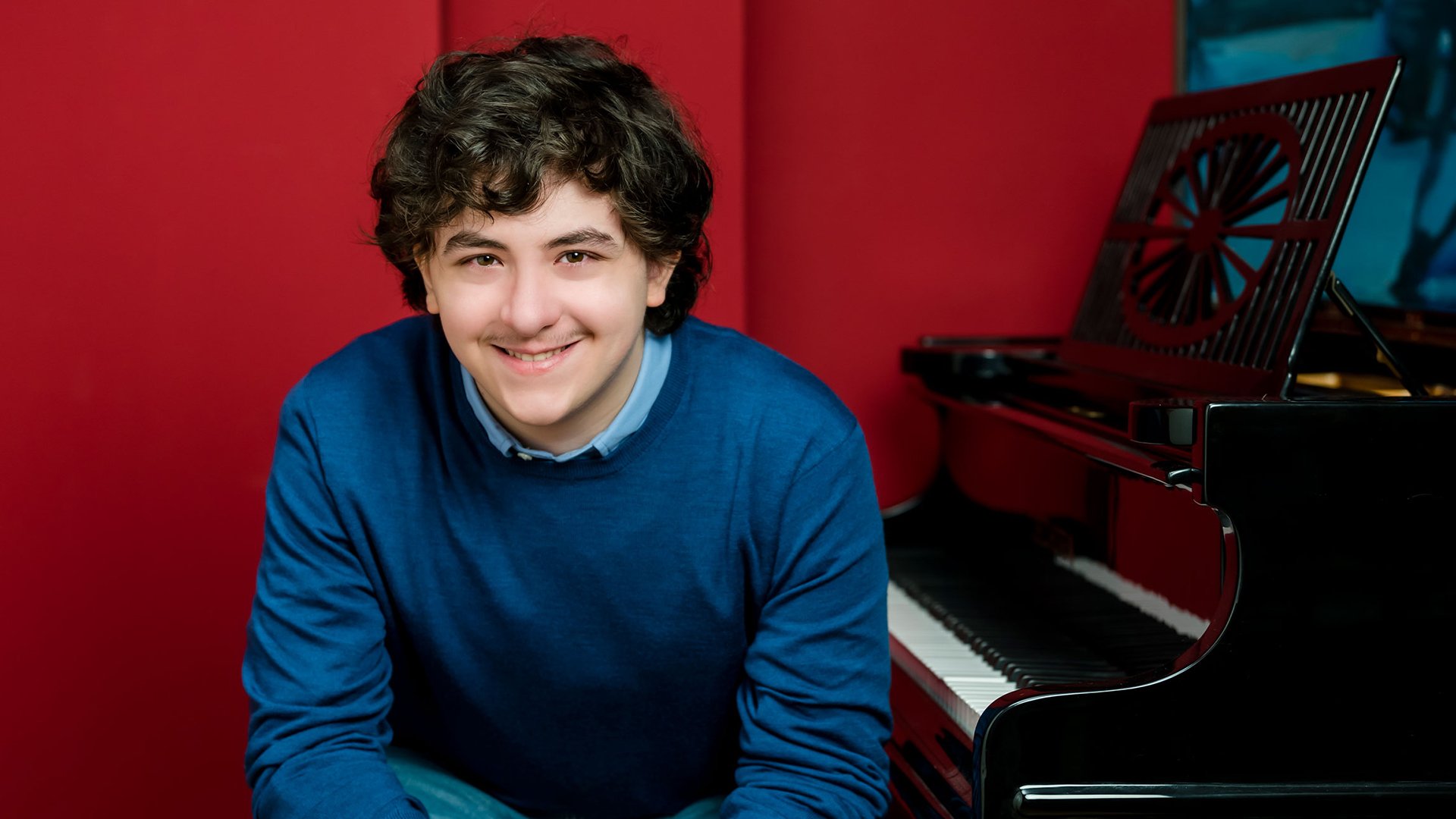 Tsotne Zedginidze in blauem Pullover an einem Piano sitzend vor rotem Hintergrund