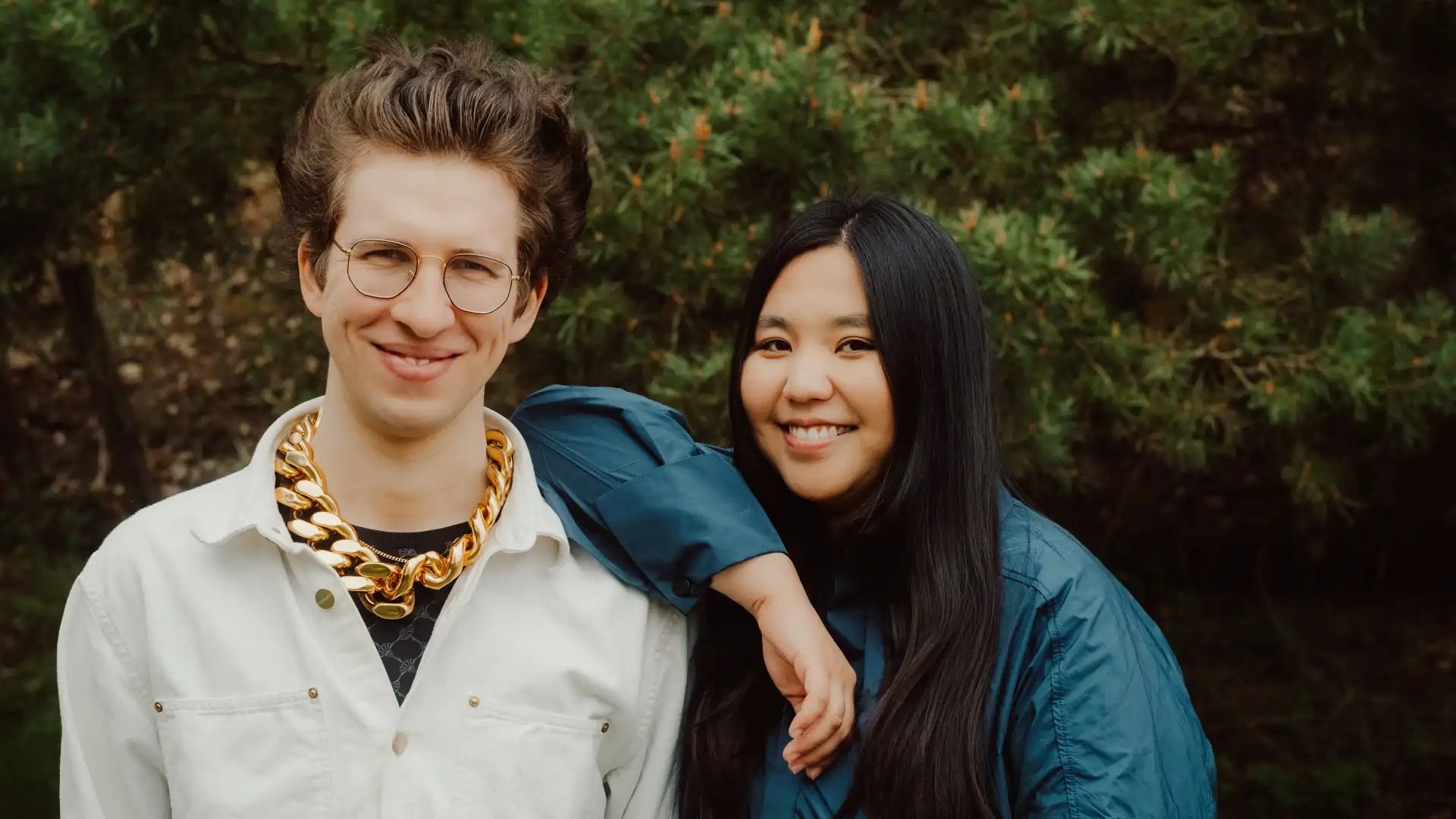 Shuteen Erdenebaatar und Nils Kugelmann vor dichtem Grün, er mit runder Brille, hellem Hemd und auffälliger goldfarbener Kette, sie in dunkelblauer Jacke mit langem Haar, beide nebeneinander in die Kamera blickend.