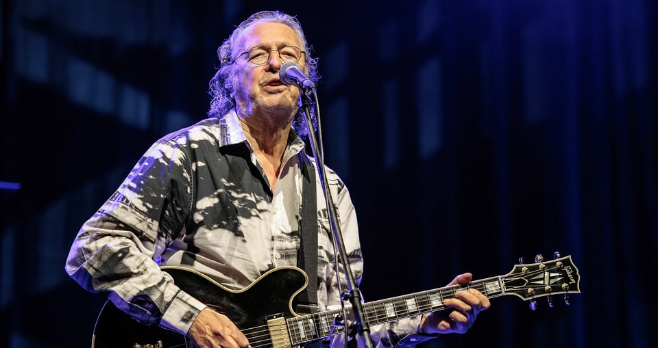 Der Künstler singt mit Mikrofon und Gitarre vor einem dunkelblauen Hintergrund.