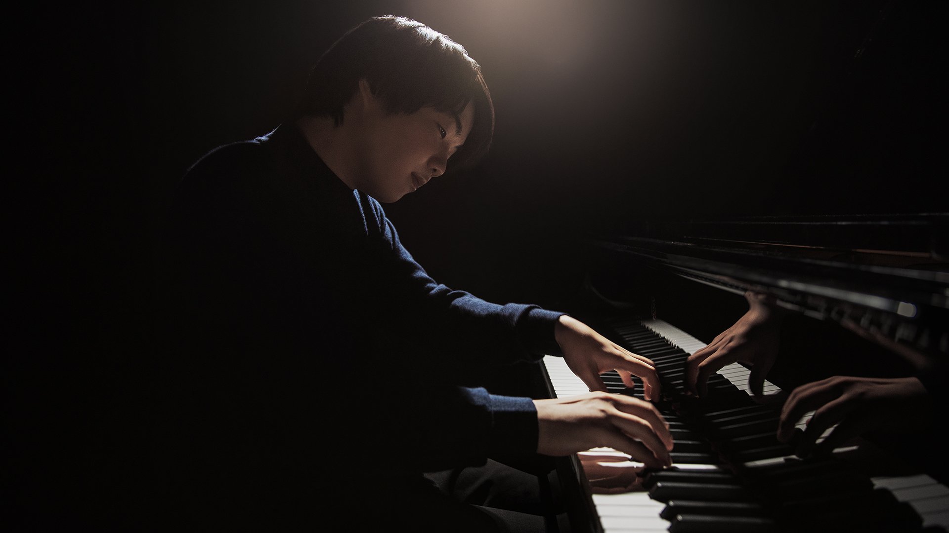 Mao Fujita spielt Klavier in dunkler Umgebung, seitlich beleuchtet mit sichtbarer Spiegelung auf dem Instrument.