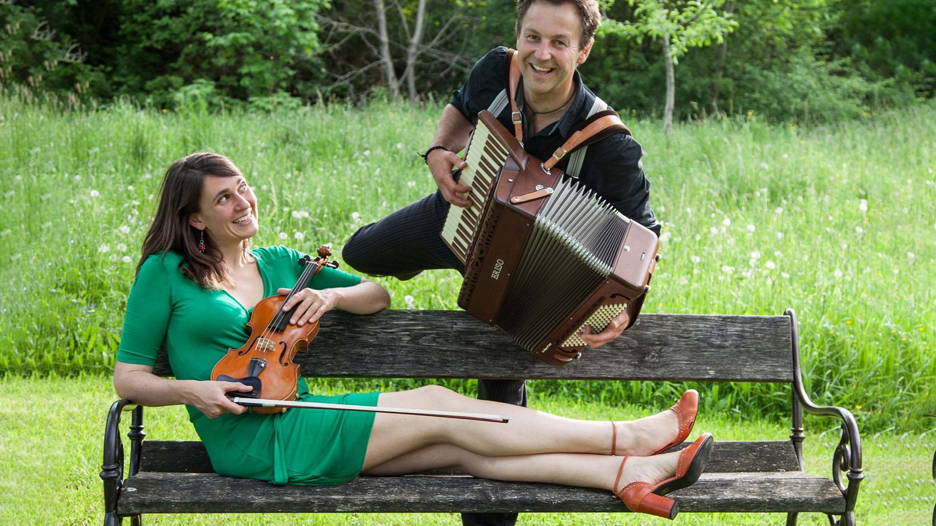 Foto eines Musikduos im Freien mit einer Person in grünem Kleid und Violine auf einer Parkbank sowie einer weiteren Person mit Akkordeon, vor einer grünen Wiese und Bäumen im Hintergrund.