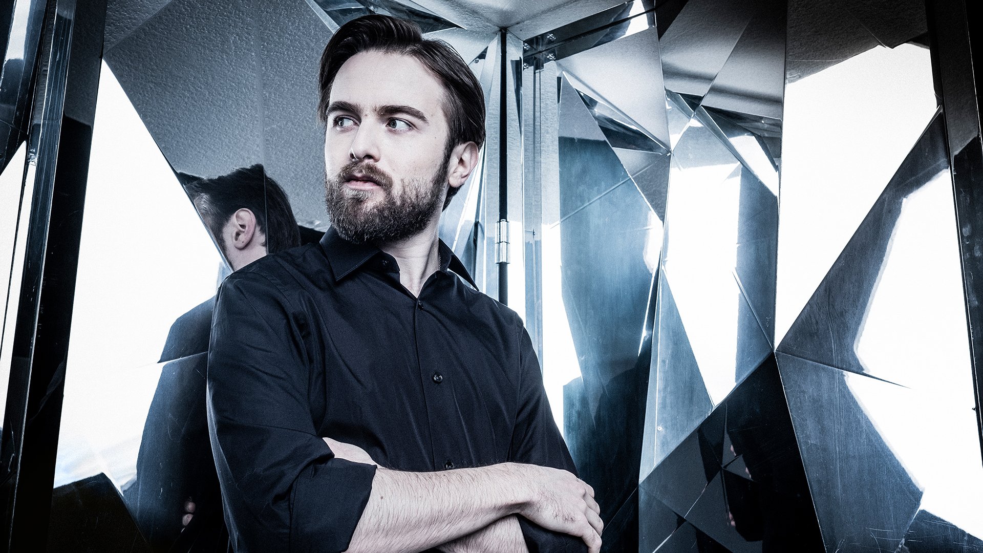Promobild für das Event ‚Daniil Trifonov, Klavier‘: Eine Person in dunklem Hemd steht mit verschränkten Armen vor einer Wand aus spiegelnden, geometrisch gebrochenen Glasflächen