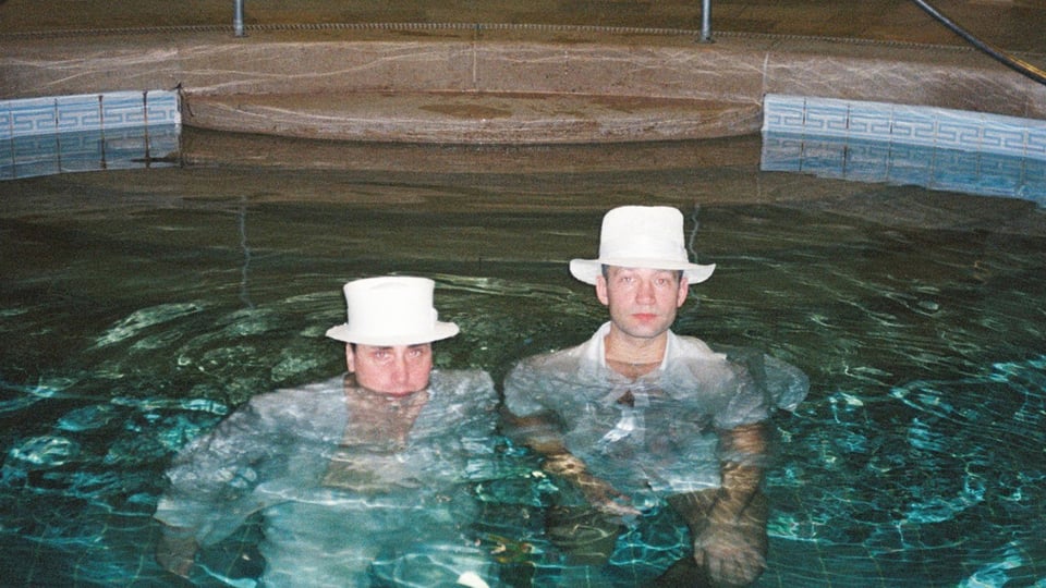 Foto für das Event „Das weiße Dorf“ mit zwei Männern, die in einem Pool stehen, beide mit weißen Hüten.