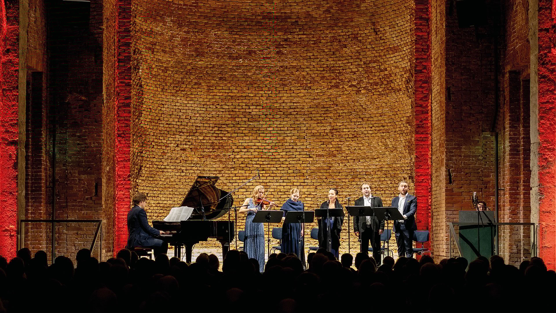 Konzertszene in der Allerheiligen-Hofkirche der Residenz München: Sängerinnen und Sänger mit Klavier und Violine bei einer klassischen Aufführung vor Backsteinwand mit roter Beleuchtung.