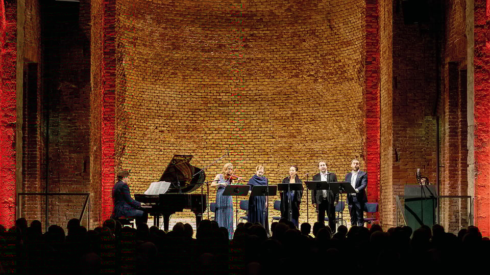 Konzertszene in der Allerheiligen-Hofkirche der Residenz München: Sängerinnen und Sänger mit Klavier und Violine bei einer klassischen Aufführung vor Backsteinwand mit roter Beleuchtung.