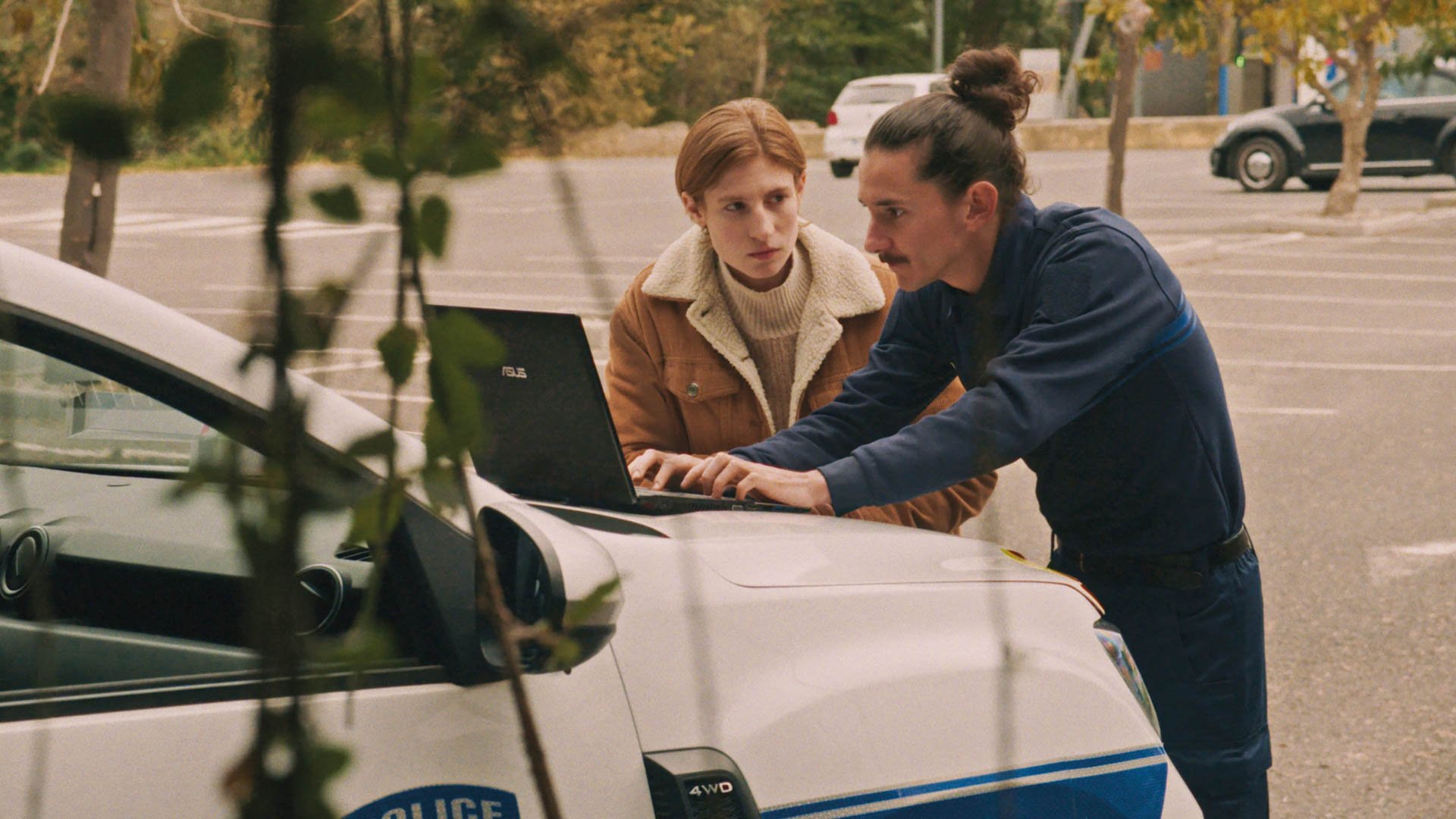 Zwei Personen stehen an einem Auto auf einem Parkplatz und schauen gemeinsam auf einen geöffneten Laptop auf der Motorhaube.
