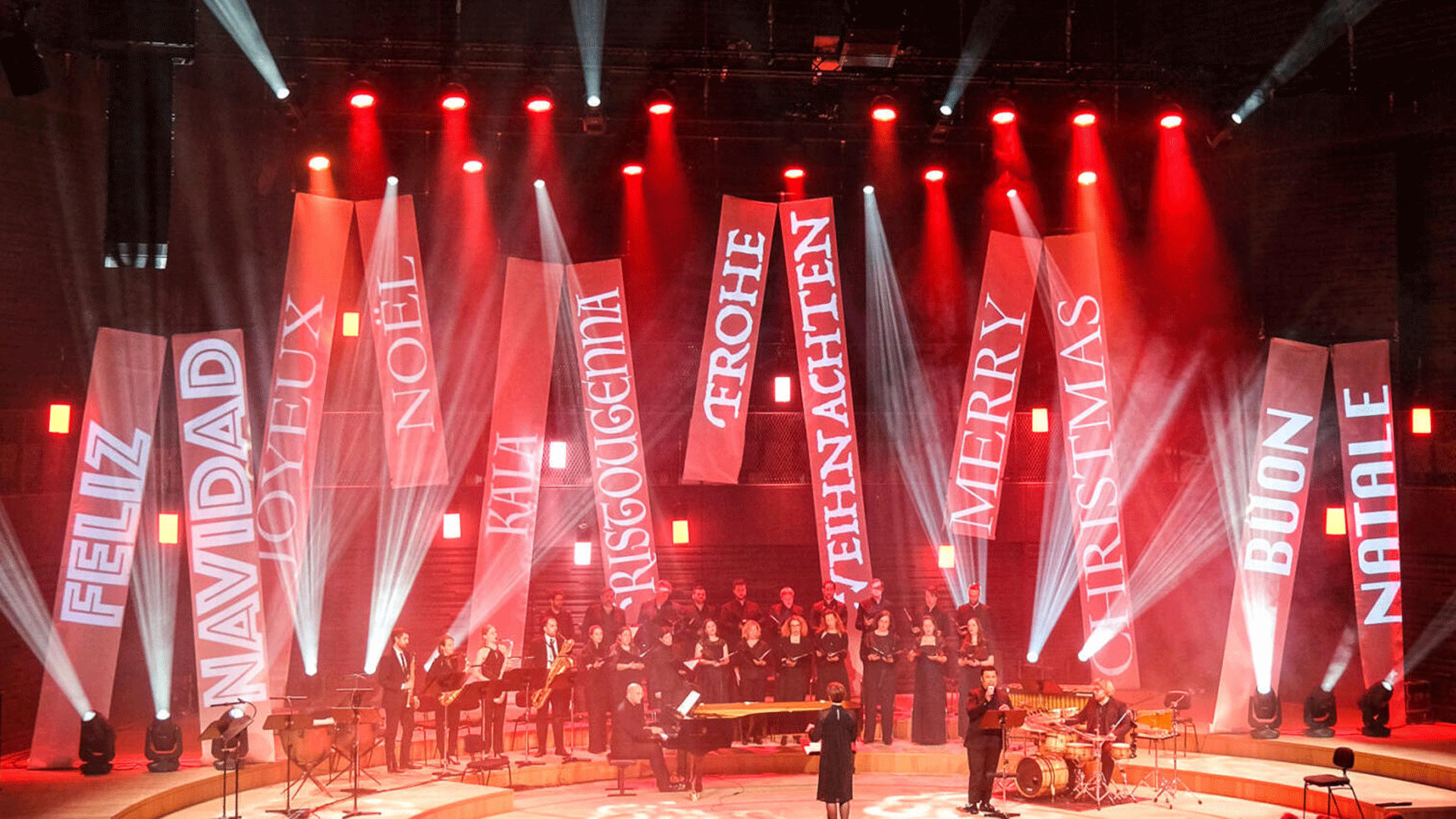Weihnachtskonzert mit Chor und Ensemble auf einer Bühne unter roter Lichtinszenierung. Hinter den Musikerinnen und Musikern hängen große Banner mit weihnachtlichen Begriffen wie Feliz Navidad, Noel, Frohe Weihnachten, Merry Christmas und Buon Natale.