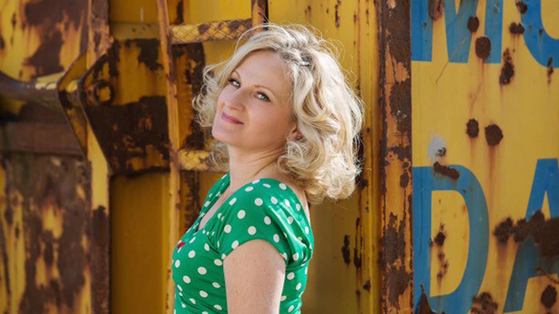 Lucia Appel steht vor einer gelben, verwitterten Metallwand und blickt seitlich zur Kamera; sie trägt ein grünes Kleid mit weißen Punkten.