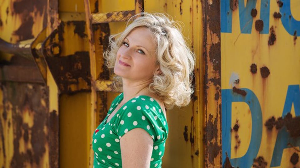 Lucia Appel steht vor einer gelben, verwitterten Metallwand und blickt seitlich zur Kamera; sie trägt ein grünes Kleid mit weißen Punkten.