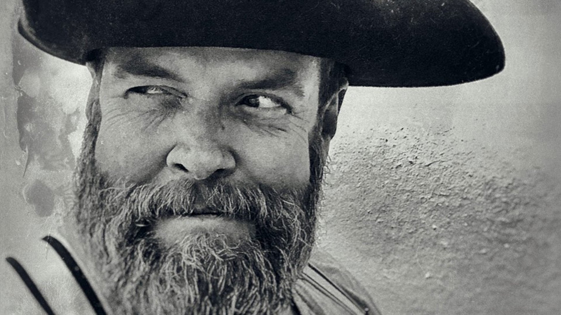 Ein schwarz-weiß Portrait von Jochen Malmsheimer mit Bart und Hut, der grinsend zur Seite blickt,  vor verschwommenem Hintergrund