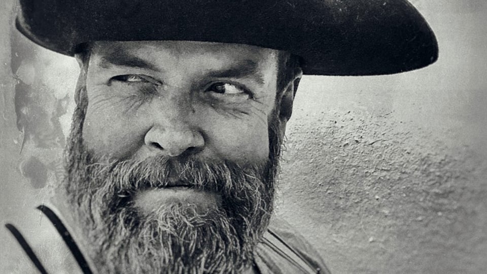 Ein schwarz-weiß Portrait von Jochen Malmsheimer mit Bart und Hut, der grinsend zur Seite blickt,  vor verschwommenem Hintergrund