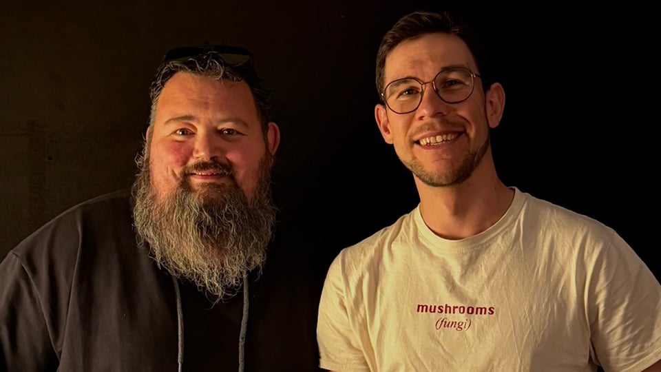 Zwei Personen vor dunklem Hintergrund; die eine trägt ein dunkles Oberteil mit langem Bart, die andere eine Brille und ein T‑Shirt mit der Aufschrift mushrooms - fungi, warm beleuchtet.