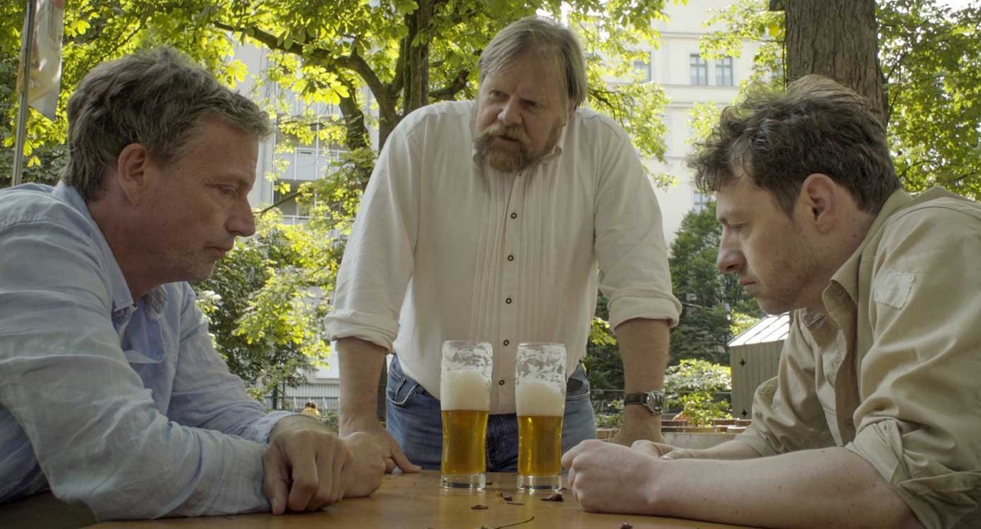 Foto von zwei Männern, die an einem Holztisch sitzen, mit jeweils zwei Bier vor sich, gegenüber voneinander, und eine dritte Person steht und schaut zu einem von ihnen.