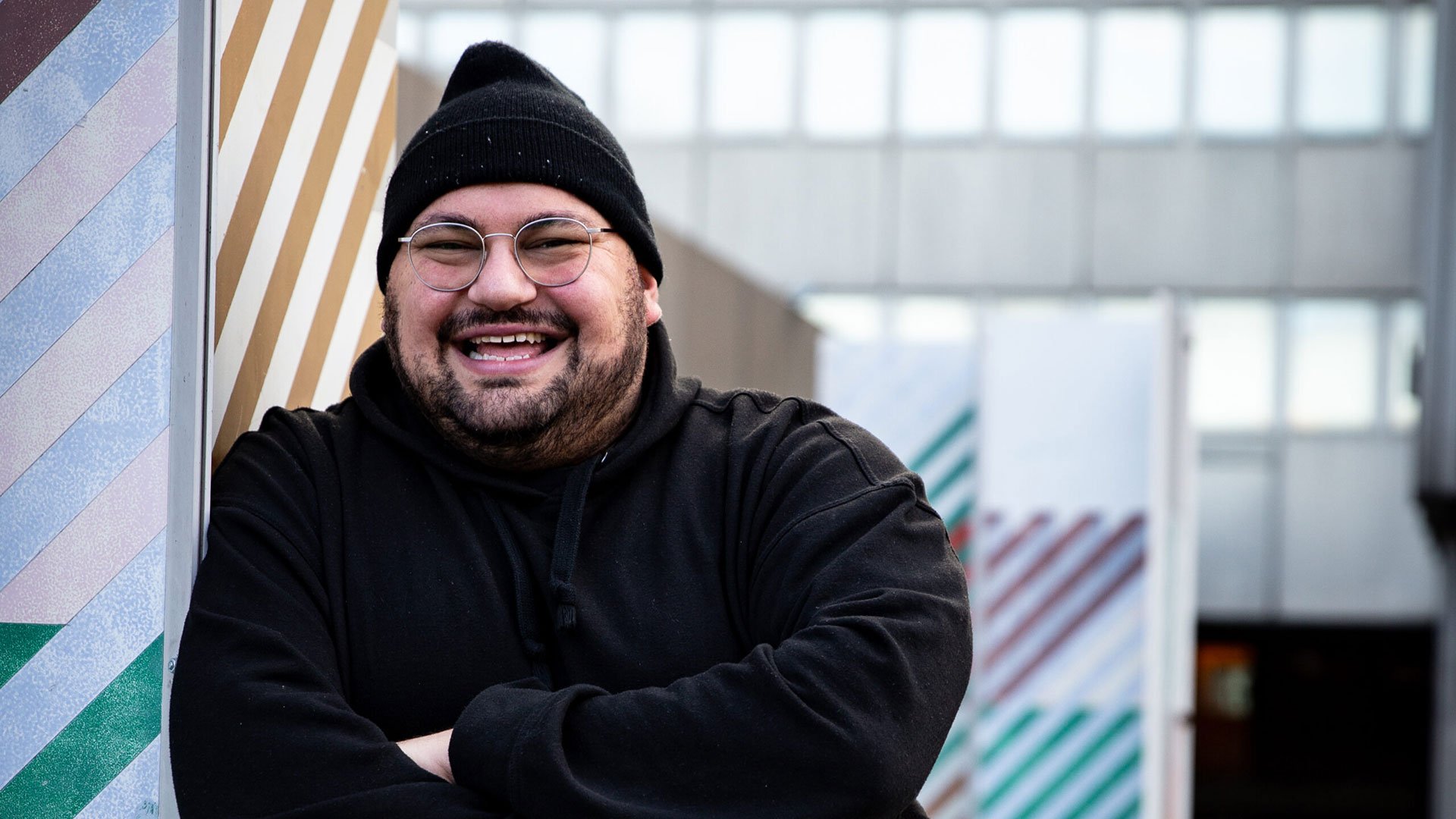 Foto für die Veranstaltung von Bora mit dem Künstler mit Sehbrille und Mütze, und im Hintergrund ein graues Gebäude mit einigen farbigen Streifen